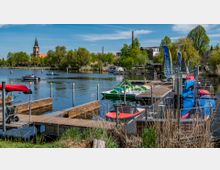 Ein kleiner Hafen mit Holzstegen, an denen Boote, Tretboote und bunte Kajaks liegen, umgeben von Schilf und einer grünen Landschaft. Im Hintergrund ist eine Kirche mit einem spitzen Turm sowie einzelne Gebäude und Bäume zu sehen.
