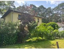Zweistöckiges Wohnhaus mit hellgelber Fassade und Satteldach, teilweise von dichtem Gartenbewuchs und einem Zaun im Vordergrund verdeckt. Im Hintergrund stehen hohe Kiefern, die das Haus in eine waldnahe Wohngegend einbetten.
