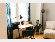 Ein hell eingerichtetes Zimmer mit einem Schreibtisch vor einem Fenster mit floralen Vorhängen. Auf dem Schreibtisch stehen eine Tischlampe, ein Buchhalter, eine Vase und eine Kerze. Daneben stehen ein grüner Stuhl, ein beiger Sessel und eine Zimmerpflanze in einem geflochtenen Korb.