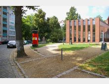 Ein kleiner Platz mit einem Weg aus Kies und Rasenflächen, umgeben von Bäumen und einem roten, runden Werbesäulen-Kiosk. Rechts steht ein modernes Gebäude mit roten Backsteinwänden und großen Glasfenstern.