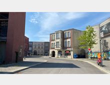 Straßenszene mit mehreren Gebäuden: Im Vordergrund ein Eckgebäude im Stil eines Altbaus mit roten und beigen Fassadenelementen, im Hintergrund weitere Gebäude, darunter ein beige-gestrichenes Haus. Eine kleine Grünfläche mit einem Baum und eine Mauer mit Graffiti runden die Umgebung ab. Klarer Himmel bei Tageslicht.