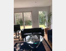 Wohnzimmer mit großen Fenstern, die auf einen Garten mit Bäumen blicken. Im Raum befinden sich ein schwarzes Ledersofa, ein runder Glastisch, ein blauer Teppich und ein Fernseher auf einem Metallgestell, daneben eine große Zimmerpflanze in einem Topf.