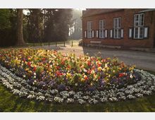 Ein historisches Backsteingebäude mit weißen Fensterläden neben einem bunt bepflanzten Blumenbeet, das von weißen Blumen umrandet ist; dahinter hohe Bäume und ein gepflasterter Weg.