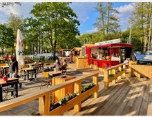 Ein Outdoor-Cafébereich mit Holzmöbeln und einer roten Imbissbude, beschriftet mit "Berlin Müggelsee", umgeben von großen Bäumen und einem nahegelegenen Segelbootanleger. Im Vordergrund befinden sich Pflanzenkästen und sonnendurchflutete Terrassenplatten.