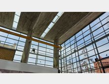 Innenraum eines modernen Gebäudes mit einer hohen Decke aus Beton und großen verglasten Wänden, die den Blick nach draußen auf Bäume und ein Backsteingebäude ermöglichen. Eine gelbe Trägerkonstruktion und künstliche Vogelaufkleber an den Fenstern sind sichtbar.