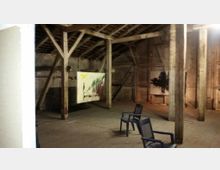 Ein rustikaler Dachbodenraum mit Holzbalken und schrägem Dach. Zwei moderne Kunstwerke sind an den Wänden aufgehängt, darunter abstrakte Malereien, und einige schwarze Kunststoffstühle stehen im Raum verteilt.