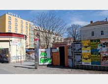 Straßenecke mit einem gelben Hotelgebäude im Hintergrund, einem orangefarbenen Fotoautomaten und einer Telefonzelle. Neben dem Automaten befinden sich Plakatwände mit Veranstaltungsankündigungen und Graffiti auf einer Betonfläche, während kahle Bäume den Ort umrahmen.