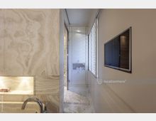 Eine moderne Badezimmeransicht mit beiger Marmorverkleidung, einer eingelassenen beleuchteten Ablage, einer verchromten Armatur und einer Glasdusche im Hintergrund. Rechts ist eine Wand mit einem flachen Fernseher und einem Fenster mit strukturierter Mattglasverkleidung.