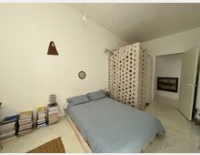 Ein minimalistisches Schlafzimmer mit einer weißen Ziegeltrennwand, einem Bett mit hellblauer Bettwäsche und einem Stapel Bücher auf dem Boden. Die Wände sind weiß, an einer hängt ein kleines, rundes Holzspiegel und ein geometrisches Kunstobjekt. Im Hintergrund ist eine geöffnete Tür zu sehen, die zu einem weiteren Raum mit einem gerahmten Bild führt.