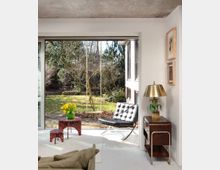 Modern living room with white walls and a concrete ceiling, featuring a large sliding glass door that opens to a backyard garden with trees and a hanging swing. Inside are a black tufted lounge chair, a small red table with yellow flowers, and a wooden side cabinet with a brass lamp and framed artwork.