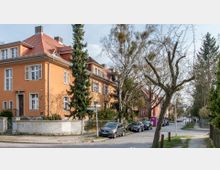 Straßenszene mit mehrstöckigen Wohnhäusern im Stil der frühen Moderne, orange gefärbten Fassaden und roten Ziegeldächern. Entlang der gepflasterten Straße stehen geparkte Autos, Bäume mit noch kahlen Ästen und Straßenschilder an einer Kreuzung.