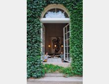 Eine offene, große Fensteröffnung eines Gebäudes, das vollständig mit grünem Efeu bewachsen ist. Dahinter sitzt ein Paar an einem gedeckten Tisch in einem hellen Raum mit hohen Decken, dekoriert mit moderner Kunst und hängenden Kugelleuchten.