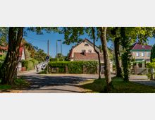 Straßenszene in einer ruhigen Wohngegend mit Einzelhäusern, umgeben von Bäumen und Hecken. Im Hintergrund führt eine kleine Brücke zu einem Fuß- und Radweg, während ein Radfahrer über den Asphaltweg fährt.