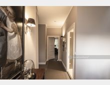 Ein heller Flur mit beige gestrichenen Wänden, einer Wandlampe, einem großen Gemälde auf der linken Seite und einer Kommode mit gläserner Dekoration. Am Ende des Flurs sieht man eine geöffnete Tür, die in einen Raum mit einem Fenster führt.