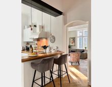 Moderne Küche mit heller Wandfarbe, einer Holz-Arbeitsfläche, zwei hohen Polsterstühlen und einer Kupferrückwand hinter dem Herd. Im Hintergrund öffnet sich ein Wohnzimmer mit großem Fenster, einem Sofa und einem gemusterten Sessel.