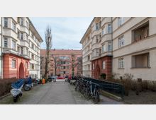 Wohnhof mit mehrstöckigen Wohngebäuden im Stil der 1920er bis 1930er Jahre, links und rechts flankiert von cremefarbenen und rötlichen Fassaden mit dekorativen Eingangsportalen. Im Vordergrund Gehwege mit abgestellten Fahrrädern, teils mit Planen bedeckt, sowie Büsche; im Hintergrund ein weiteres Gebäude mit orangefarbener Fassade.