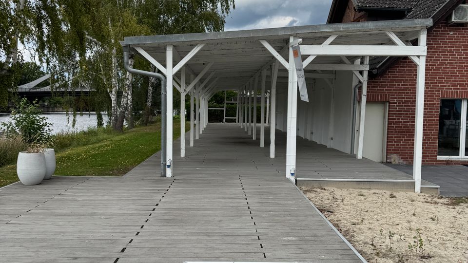 Ein langer überdachter Holzsteg führt vom rechten Rand eines Backsteingebäudes mit dunkelgrauem Dach entlang einer Wiese und eines sandigen Bereichs. Im Hintergrund sind Bäume und ein Fluss zu sehen, unter einem bewölkten Himmel.