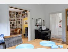 Esszimmer mit hellem Holztisch und blauen Platzdeckchen im Vordergrund. Im Hintergrund führt eine offene Tür zu einem Wohnraum mit großen, eingebauten Bücherregalen und einem braunen Sofa. Rechts im Bild steht ein schwarzes Klavier an der Wand.