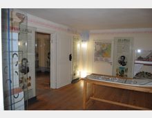 Ein kleiner Museumsraum mit Holzboden und hell gestrichenen Wänden. Entlang der Wände befinden sich Informationspaneele, darunter Landkarten und historische Bilder, sowie ein Modell eines Gebäudes und ein Ausstellungstisch in der Mitte.