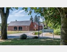 Ansicht einer ländlichen Straße mit einem Wegweiser zur "Elbtalklinik" auf der rechten Seite. Im Hintergrund befinden sich rot verklinkerte Gebäude mit Satteldächern, umgeben von Bäumen und Grünflächen.