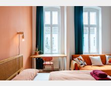 Schlafen-Wohnbereich mit einem Doppelbett, einer orangefarbenen Couch, einem Holztisch vor zwei großen Fenstern mit blauen Vorhängen und einer rosa Stuhl. Eine Topfpflanze steht auf dem Tisch, und im Hintergrund ist ein Ausblick auf Nachbargebäude zu sehen.