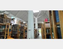 Innenraum einer modernen Bibliothek mit hohen Decken und sichtbarer Dachkonstruktion. Regale aus hellem Holz, gefüllt mit Büchern, erstrecken sich über zwei Etagen; Tageslicht fällt durch große Fensterelemente ein.