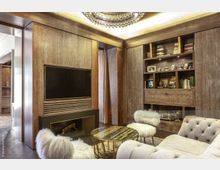 Hochwertig gestaltetes Wohnzimmer mit Holzwänden, integriertem Flachbildschirm, einem Regal mit Büchern und Bildern sowie luxuriösen Möbeln wie gepolsterten Sesseln mit flauschiger Oberfläche und einem runden Couchtisch mit Marmoreffekt. Oben hängt ein großer, dekorativer Kronleuchter an einer abgehängten Decke.