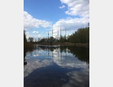Stillgewässer in einer natürlichen Umgebung mit Schilf und Bäumen am Ufer, im Hintergrund mehrere hohe Funkmasten und ein teilweise bewölkter Himmel, der sich im Wasser spiegelt.