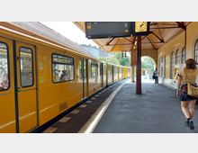 Gelber U-Bahn-Zug an einem perronüberdachten Bahnsteig mit Holzdach und Rundbogenfenstern; eine Uhr und ein Anzeigebildschirm zeigen die Linie U3. Passanten gehen am Bahnsteig entlang, einer nutzt einen Fahrkartenautomaten.