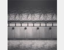 Schwarz-weiß-Aufnahme eines leeren, industriellen Innenraums mit hohen Wänden. An der Decke hängen zahlreiche lose Kabel, darunter eine Reihe kleiner, runder Fenster mit metallischen Rahmen.