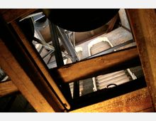 Blick von unten in das Innere eines Glockenturms mit Holzbalken, Stahlträgern und Glockenmechanismus, umgeben von gemauerten Wänden mit schmalen Fenstern und dämmendem Lichteinfall.