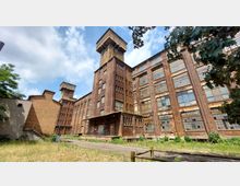 Ehemaliges Industriegebäude aus Backstein mit großen, teilweise beschädigten Fenstern und einem hohen Turm in der Mitte. Im Vordergrund befinden sich Grasbewuchs, ein unbefestigter Weg und Absperrungen.