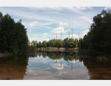 Ein ruhiger See mit klarem Wasser, umgeben von Bäumen und Schilf. Im Hintergrund sind mehrere hohe Funkmasten zu sehen, eingebettet in eine Landschaft mit blauem Himmel und Wolken.