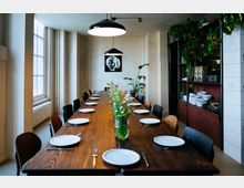 Langer Holztisch in einem modernen, minimalistisch gestalteten Essbereich, gedeckt mit weißen Tellern und Besteck, dekoriert mit Blumen in Vasen; im Hintergrund sind hohe Fenster, Pflanzen und ein Regal mit Geschirr sichtbar. Zwei große, schwarze Hängelampen bieten Beleuchtung, und an der Wand hängt ein gerahmtes Bild.