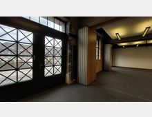 Innenansicht eines Raumes mit industriellem Design; links eine große Glastür mit einem Gittermuster, die nach draußen zu einem Wasserkanal und Backsteingebäuden führt, rechts ein Raumteiler aus Holzlamellen unter einer beleuchteten Decke.