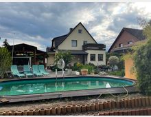 Wohnhaus mit Satteldach und angebauter Terrasse, umgeben von einem Garten. Im Vordergrund befindet sich ein rechteckiger Swimmingpool mit Holzumrandung, daneben mehrere Liegestühle.