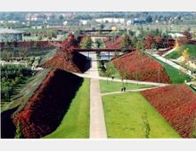 Weitläufiger Park mit symmetrisch angelegten, grasbedeckten Wegen und bepflanzten Böschungen in Rot- und Grüntönen. Im Hintergrund sind Brücken, Bäume und Wohnhäuser sichtbar.