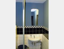 Kleines Badezimmer mit blau gestrichenen Wänden und schwarzer sowie weißer Kachelverkleidung. Ein Waschbecken mit Spiegel darüber ist an der Wand montiert, darunter sichtbare Rohrleitungen; daneben steht ein Eimer.