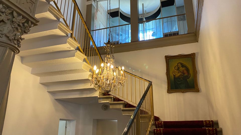 Innenansicht eines historischen Treppenhauses mit weißer, geschwungener Treppe, goldfarbenem Geländer und rotem Teppichläufer. Ein Kristalllüster hängt im Luftraum, darüber sind große Rundbogenfenster und an der Wand ein gerahmtes Gemälde zu sehen.
