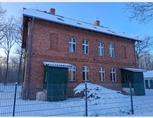 Backsteingebäude mit zwei Stockwerken, symmetrischen Fenstern und schneebedecktem Satteldach, umgeben von einem Zaun und schneebedeckter Landschaft; im Hintergrund sind Bäume eines Waldes zu sehen.