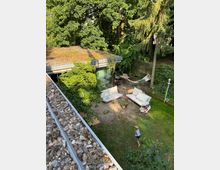 Backyard patio beside a modern, flat-roofed house with large glass doors, outdoor sofas and chairs on a sandy terrace, and a hammock near a wooden fence. The garden is bordered by tall, dense trees and shrubs, with a small lawn area in the foreground.