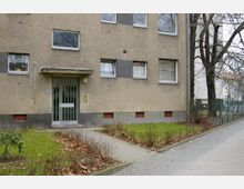 Mehrfamilienhaus mit grauer Fassade und mehreren rechteckigen Fenstern. Vor dem Gebäude befindet sich ein kleiner gepflasterter Weg, umgeben von einer Grünfläche mit kahlen Büschen und Bäumen.