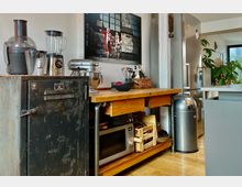 Küchenbereich mit einer Arbeitsfläche aus Holz, auf der Mixer, Rührgerät und andere Küchenutensilien stehen. Daneben ein schwarzer Metallschrank, darunter eine Mikrowelle, und im Raum befinden sich ein Edelstahlkühlschrank, eine silberne Mülltonne sowie grüne Pflanzen im Hintergrund.