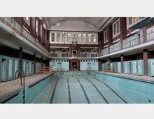 Alter Innenraum eines Schwimmbads mit leerem Becken und türkisfarbenen Fliesen. Der Raum hat hohe Decken, umlaufende Galerien mit Geländern und Türen zu Umkleidekabinen sowie große Fenster an den Wänden.