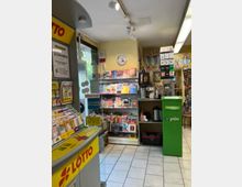 Innenraum eines Zeitschriften- und Lottogeschäfts mit Regalen voller Magazine und Zeitungen. Links steht ein gelber Lotto-Schalter, im Hintergrund befindet sich eine grüne Paketstation sowie Kaffeeautomaten und weitere Verkaufswaren.