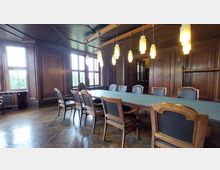 Holzgetäfelter Konferenzraum mit einem langen Tisch, der mit einer grünen Oberfläche bedeckt ist, umgeben von gepolsterten Stühlen. Große Fenster lassen natürliches Licht herein, und mehrere hängende Lampen beleuchten den Raum.