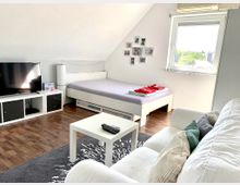 Wohnzimmer in einem Dachgeschoss mit heller Einrichtung: Ein Raum mit einer weißen Couch, Couchtisch, Flachbildfernseher auf einem Regal, einem Bett mit Bettwäsche und Handtüchern, sowie einem Fenster, das Tageslicht hereinlässt. Der Boden ist mit Holzlaminat ausgelegt, ergänzt durch einen Teppich.