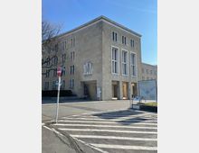 Eckgebäude eines historischen Gebäudekomplexes in hellbeigem Stein mit schlichten Fassaden und großen vertikalen Fenstern. Im Vordergrund ein Zebrastreifen, eine Informationstafel mit Aufschrift „Flughafen Tempelhof“ und karge Vegetation.