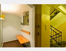 Kleiner Raum mit einer hölzernen Sitzbank an der Wand und einem kleinen Bogenfenster mit Lüftungsklappen. Rechts davon befindet sich ein Treppenhaus mit gelb gestrichenen Wänden und einem schwarzen Geländer.