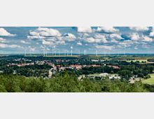 Weitläufige Landschaftsansicht mit einer kleinen Stadt im Vordergrund, umgeben von grünen Feldern und Wäldern, im Hintergrund eine Reihe von Windrädern unter einem Himmel mit vielen Wolken.